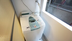 A compact, white washing machine with a control panel, located in a corner of a room with a window. - Apartment Camon EBR0001E (Osaka)