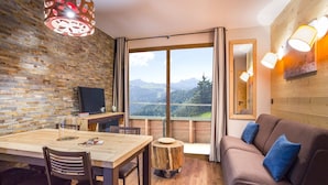 A cozy room with a wooden dining table, chairs, a sofa with blue cushions, a TV, and a large window offering a mountain view. - Alpine Stay w/ Spa + Views (Tignes 1800)