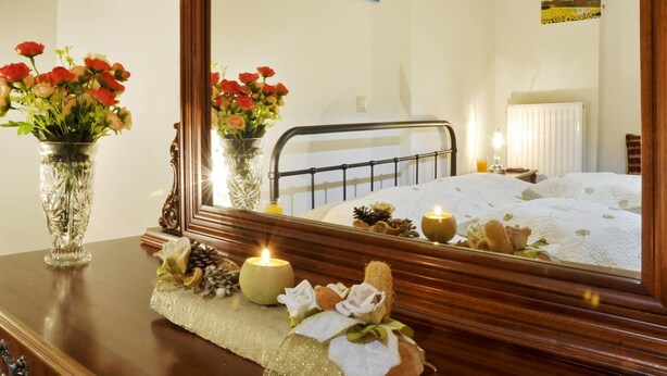 Un dormitorio con un espejo grande, una cama con ropa de cama blanca, una mesita de noche con una lámpara y dos jarrones de flores sobre un aparador.
