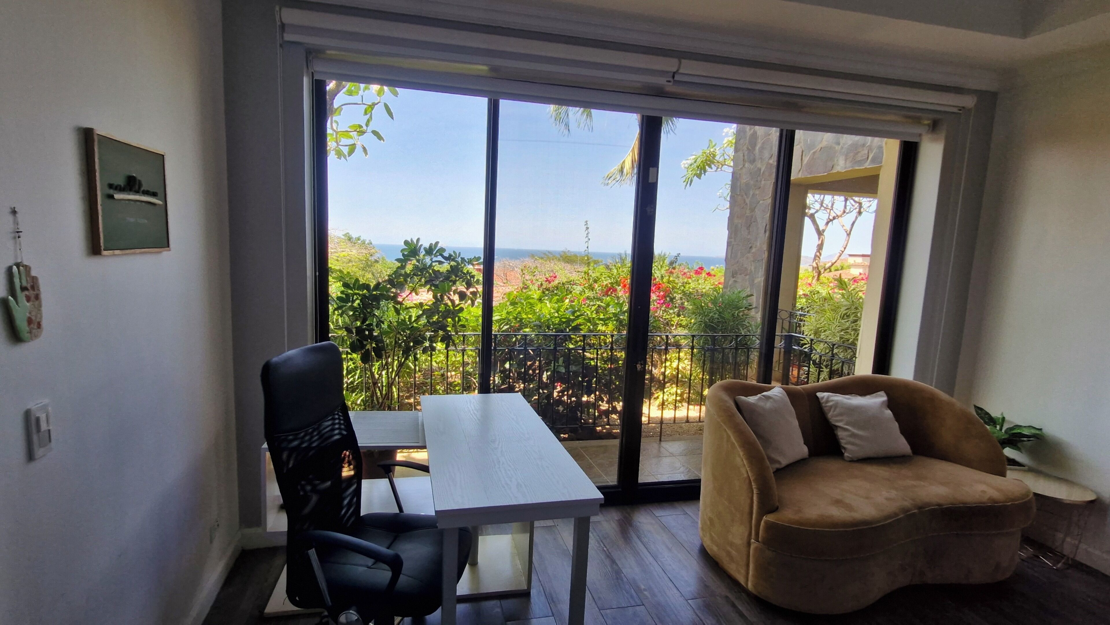 Una habitación con una mesa blanca, una silla de escritorio negra, un sofá marrón y vistas a la vegetación y al mar a través de amplios ventanales correderos.