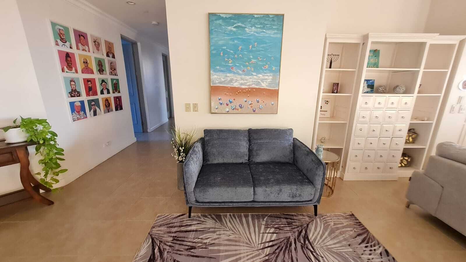 Un salón con un sofá gris, un tapete estampado, una pared con un cuadro de temática playera y un mueble blanco con cajones.