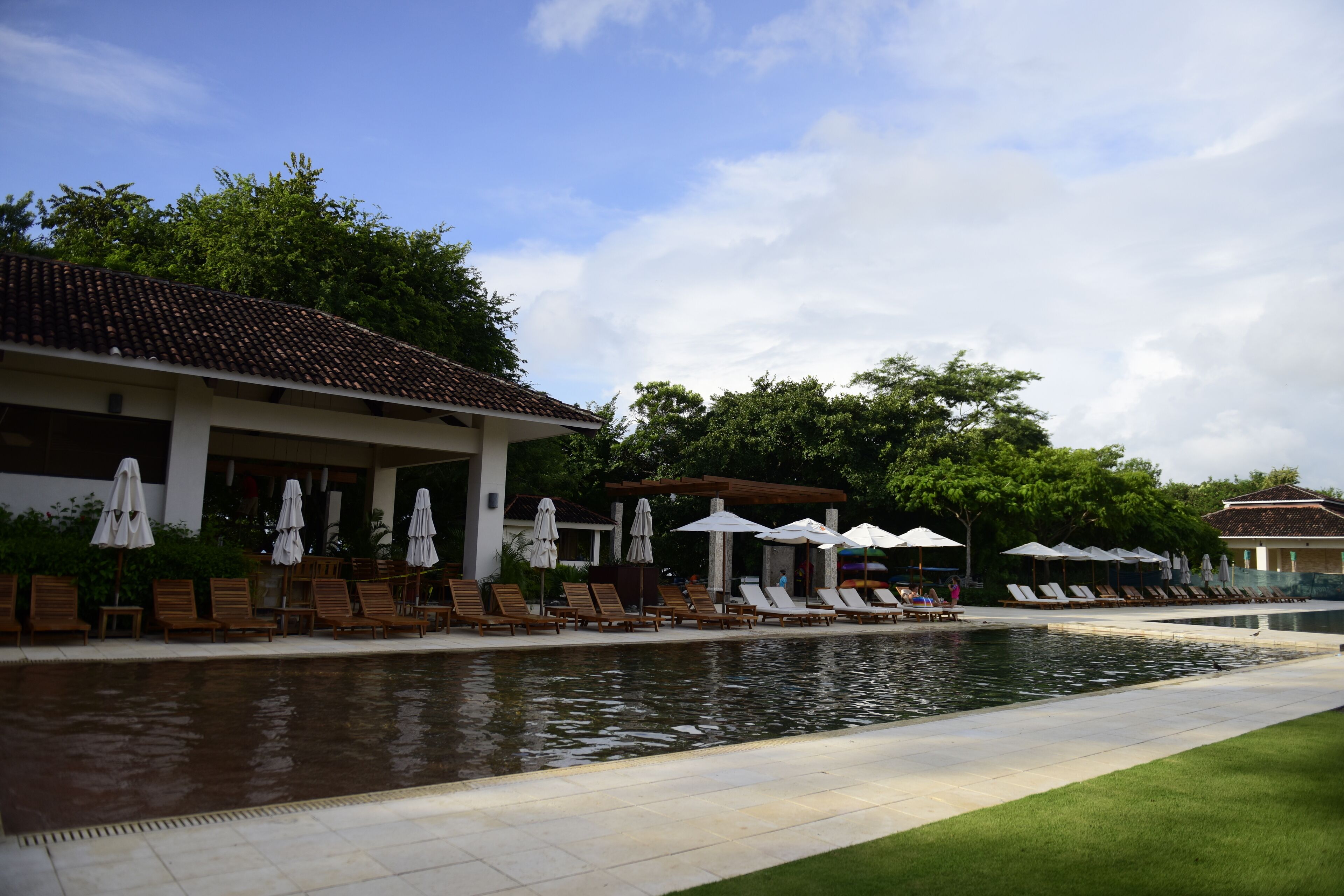A pool area with lounge chairs and umbrellas, surrounded by greenery and a building with a tiled roof.