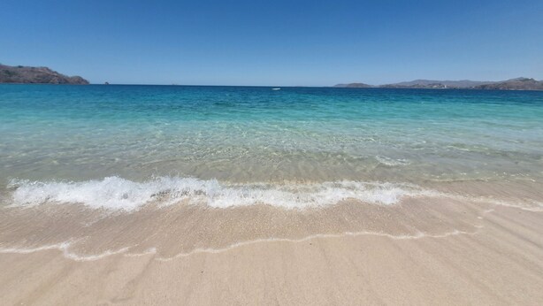 Ein Strand mit klarem Wasser, sandigem Ufer und in der Ferne Hügeln.