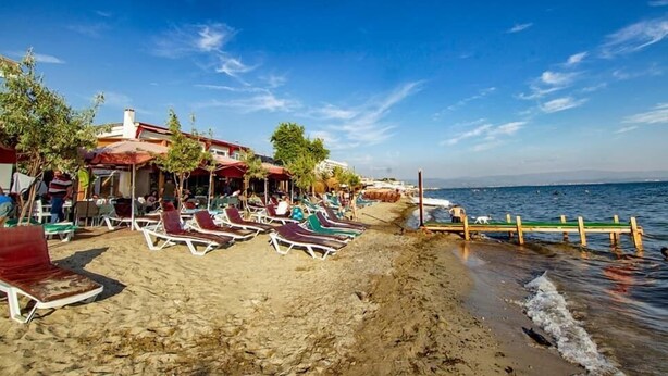A beachside area with sun loungers and umbrellas, a wooden pier extending into the water, and a building with a red roof.