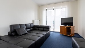 A modern living room with a sectional sofa, a flat-screen TV on a wooden cabinet, and a large window with sheer curtains. - Fly Inn Hotel Frankfurt Airport Rüsselsheim ehemals Trip Inn Hotels (Ruesselsheim)