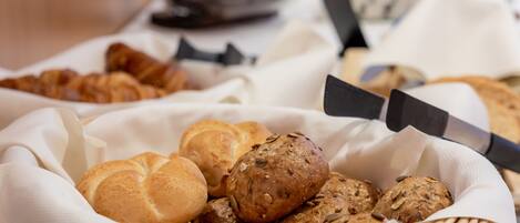 A basket of assorted breads and rolls, including croissants and seeded rolls, on a table with a knife and a tray of condiments in the background.