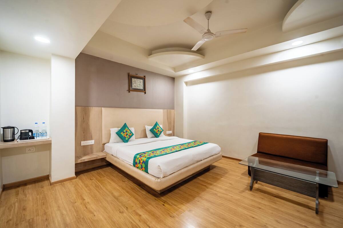 A modern hotel room with a large bed, a wooden desk with a kettle, a bench, and a ceiling fan.