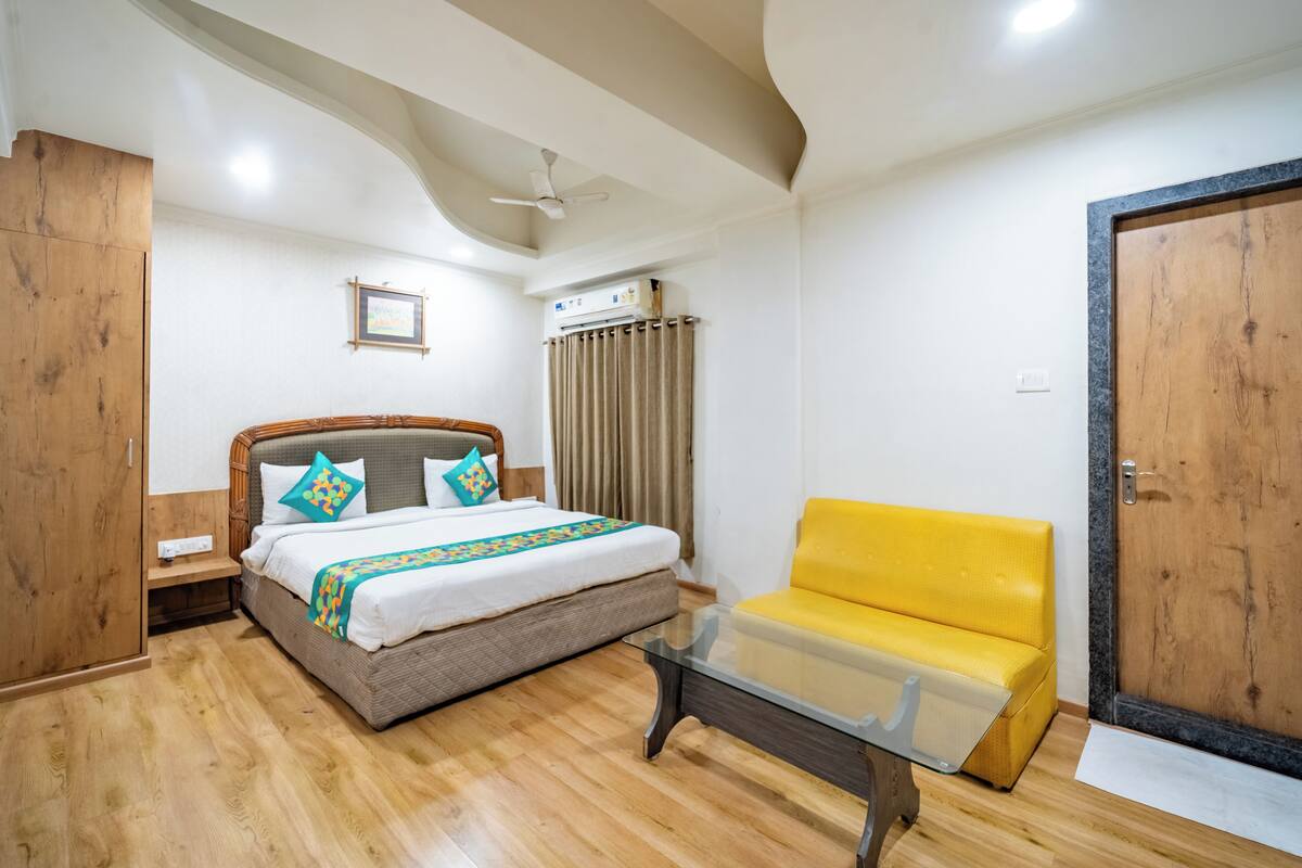 A bedroom with a bed, a yellow sofa, a glass coffee table, a ceiling fan, and a wooden door.
