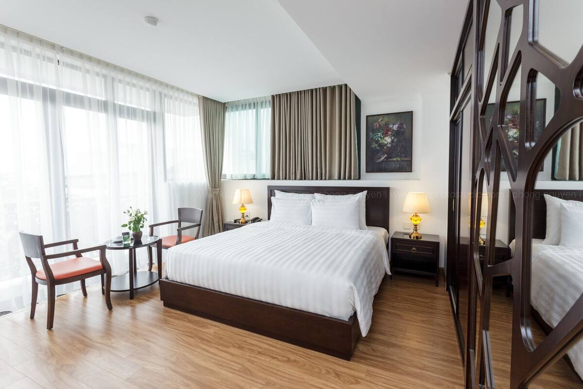 Ein Hotelzimmer mit einem großen Bett, einem Schreibtisch mit Stuhl, einem Nachttisch mit Lampe und einem Fenster mit Vorhängen.