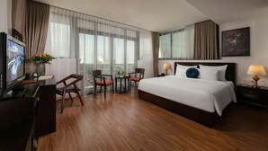 Una habitació d'hotel amb un llit gran, una taula de treball amb un televisor, dues cadires, una taula petita amb un vas i una finestra amb cortines.