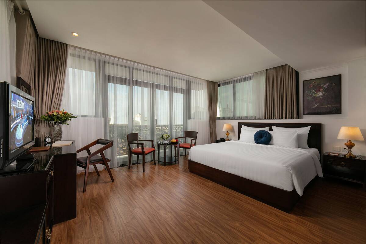 Ein Hotelzimmer mit einem großen Bett, einem Schreibtisch mit Fernseher, zwei Sesseln, einem kleinen Tisch mit Vase und einem Fenster mit Vorhängen.