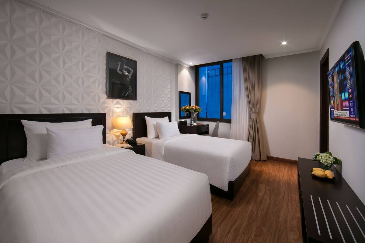 Ein Hotelzimmer mit zwei Betten, einem Nachttisch mit Lampe, einem Schminktisch, einem großen Fenster mit Vorhängen und einem an der Wand befestigten Fernseher.
