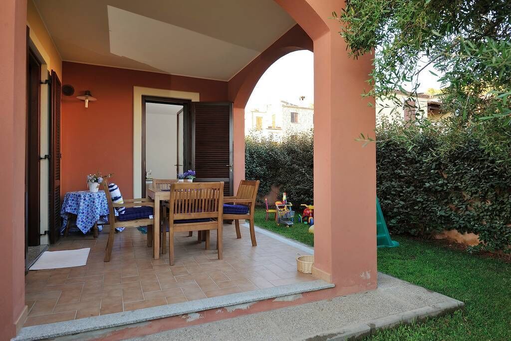 A covered patio area with a table and chairs, a door, and a view of the surrounding garden and buildings.