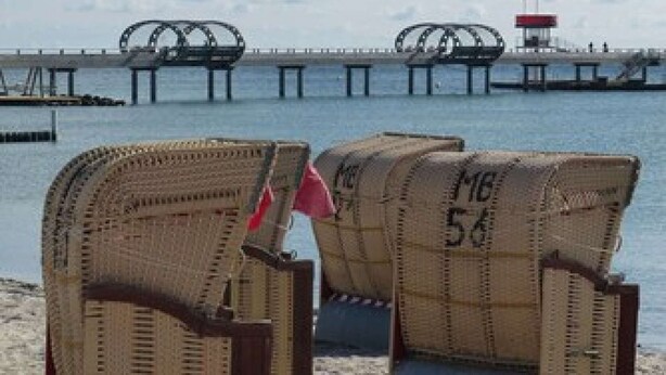 Two wicker chairs with numbers on them on a sandy beach, with a pier and a lighthouse in the background.
