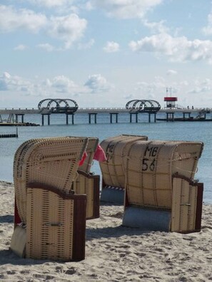 Two wicker chairs with numbers on them on a sandy beach, with a pier and a lighthouse in the background.