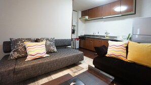 Un salon avec un canapé et une table basse, une cuisine avec des meubles en bois et un plan de travail, et un mur blanc avec un luminaire au plafond.