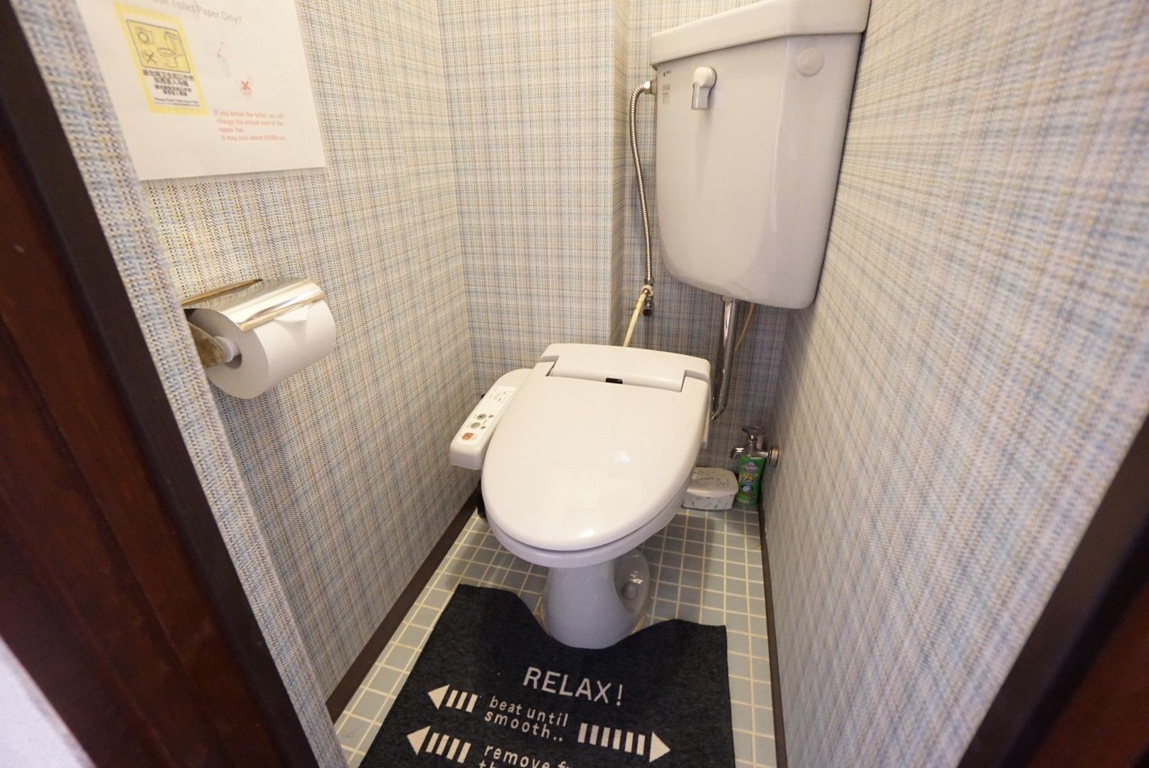 A small bathroom with a toilet, a paper towel dispenser, and a hand dryer. There is a sign on the wall and a mat on the floor with directional arrows.