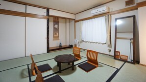 Une pièce de style japonais traditionnel, avec un sol recouvert de tatamis, une table en tatami, quatre chaises en bois, une petite étagère avec un vase, un climatiseur fixé au mur et un miroir.
