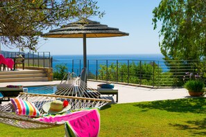 A hammock with a watermelon-shaped pillow, a striped pillow, and a straw hat, set up under a thatched umbrella on a patio overlooking a pool and the sea. - ELOUNDA  LUXURY VILLA WITH PRIVATE POOL (Elounda)