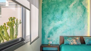 Een slaapkamer met een bed, nachtkastje, raam met uitzicht op cactussen en een muur in teal kleur.
