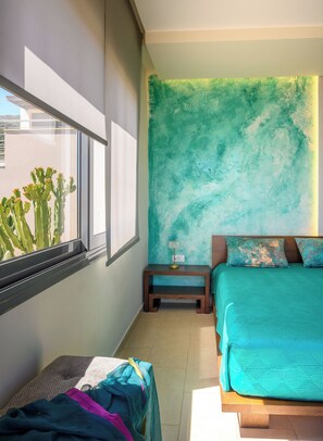 A bedroom with a bed, a nightstand, a window with a view of cacti, and a teal-colored wall.