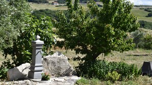 Un monument de pierre surmonté d’un pinacle, entouré de verdure et avec un paysage rural en arrière-plan.