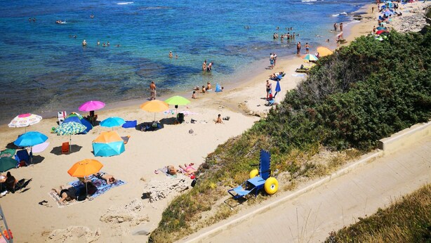 Une plage avec une eau d’un bleu limpide, des gens qui se baignent et prennent des bains de soleil, et des parasols aux couleurs vives.