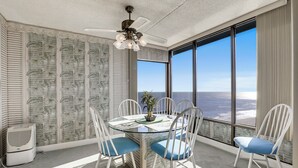 Ein Zimmer mit Glastisch, weißen Stühlen, einem Deckenventilator, einer Topfpflanze und einem großen Fenster mit Aussicht.