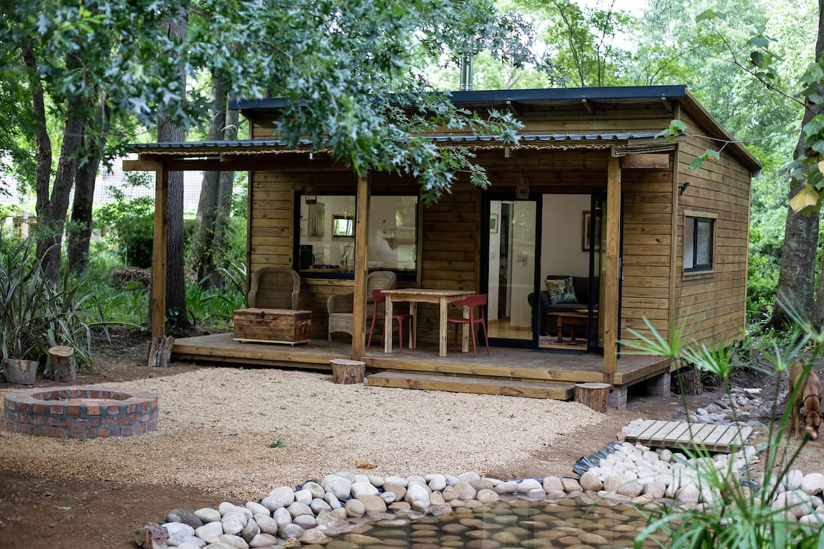 A wooden cabin with a porch, a brick fire pit, and a small pond.
