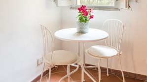 Een kleine kamer met een ronde tafel, twee witte stoelen, een raam met open luiken en een bloempot met rode bloemen.