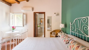 Een slaapkamer met een bed, een raam, een stoel, een tafel met een bloevas, en een badkamer zichtbaar door een open deur.