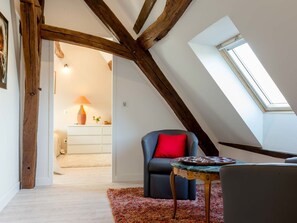 Une pièce avec des poutres apparentes, une lucarne, un fauteuil bleu foncé agrémenté d’un coussin rouge, une petite table avec un plateau et un tapis coloré.