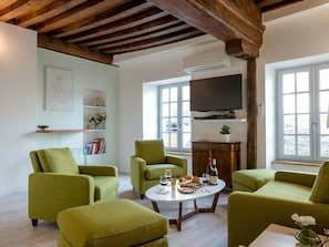 A living room with green armchairs, a round coffee table with wine glasses and food, a flat-screen TV, and a bookshelf.