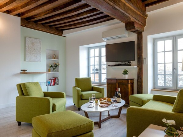 A living room with green armchairs, a round coffee table with wine glasses and food, a flat-screen TV, and a bookshelf.