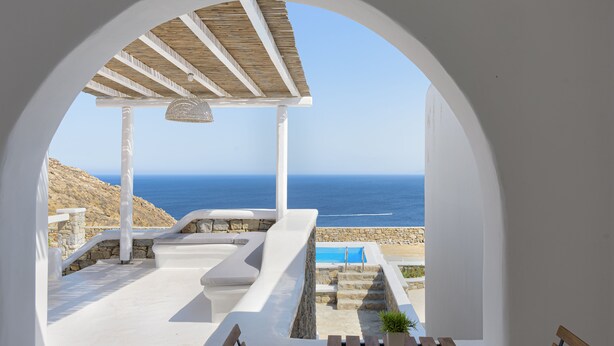 A view through an arched doorway to a pool area with a stone wall, wooden deck, and a bench. - Mykonos Unique Villas (Mykonos)
