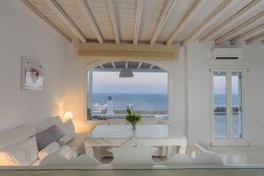 A room with a white sofa, a table with a vase, and a view of the sea through a window. - Mykonos Unique Villas (Mykonos)