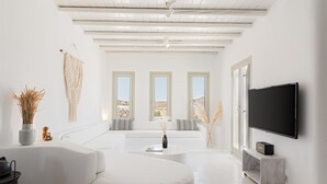 A modern living room with a white sofa, a coffee table, a flat-screen TV, and a large window with a view of the desert. - Mykonos Unique Villas (Mykonos)
