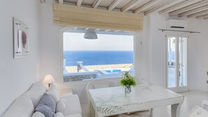 A room with a view of the sea, a white table with a plant, a sofa, and a hanging light. - Mykonos Unique Villas (Mykonos)