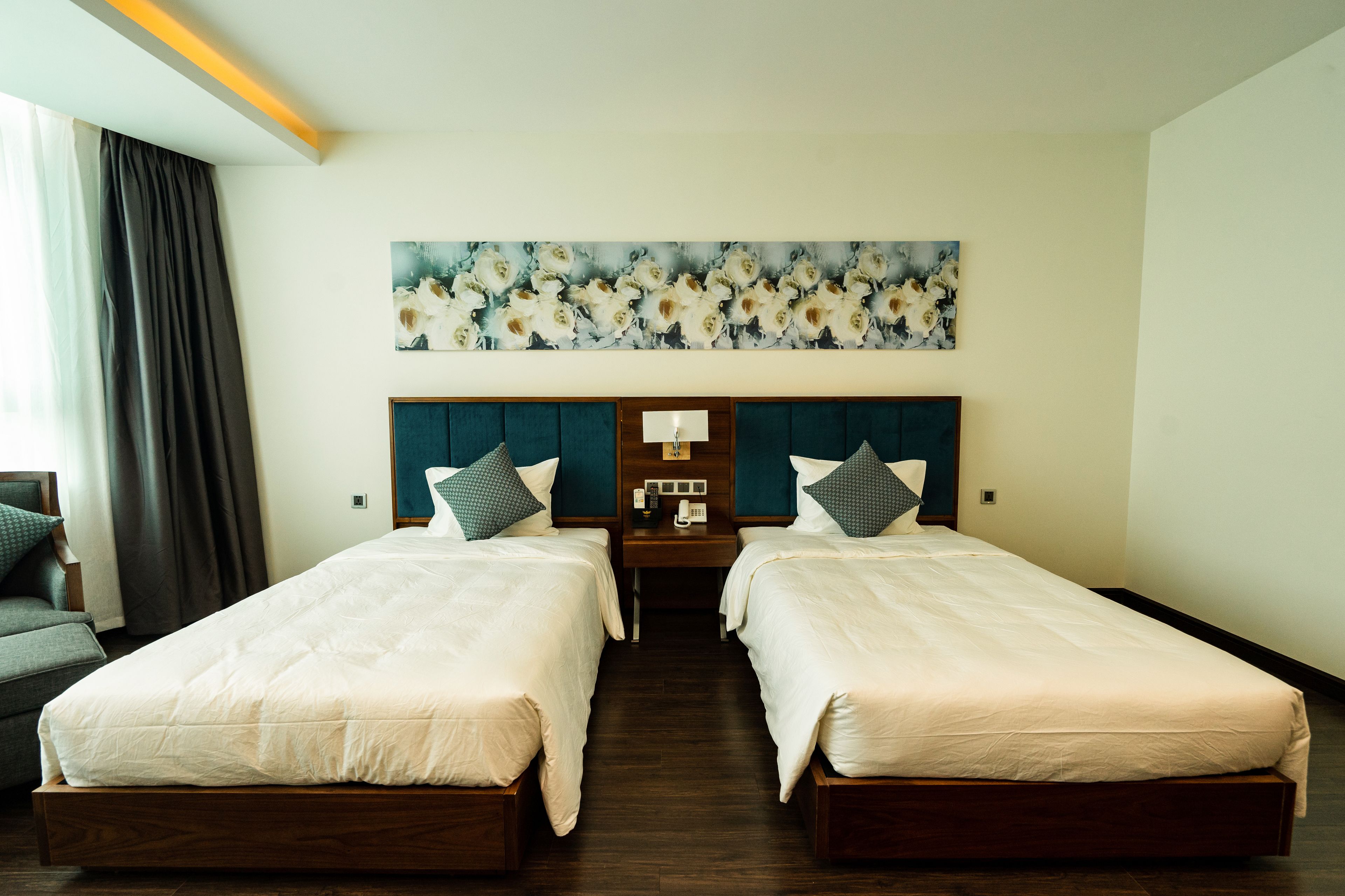 A hotel room with two beds, a nightstand, a lamp, and a large floral artwork on the wall.