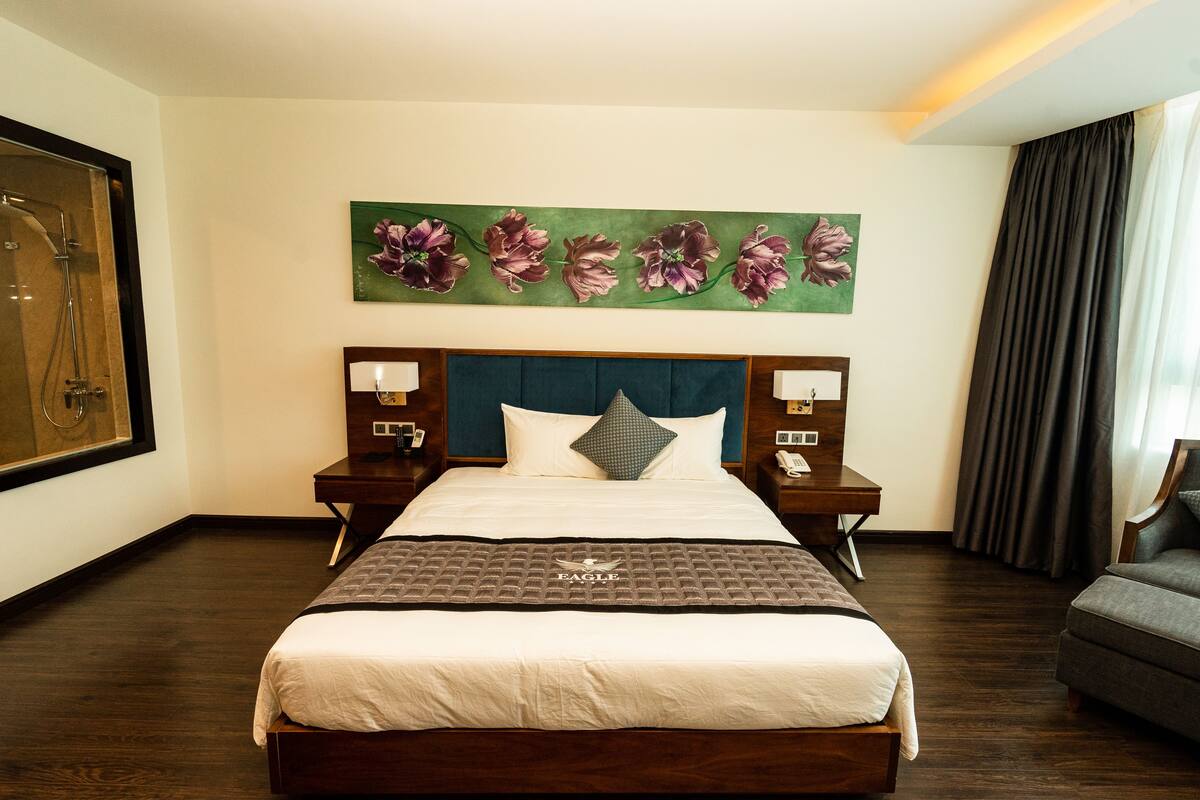 A hotel room with a large bed, a headboard with a floral painting, two bedside tables with lamps, and a grey armchair.