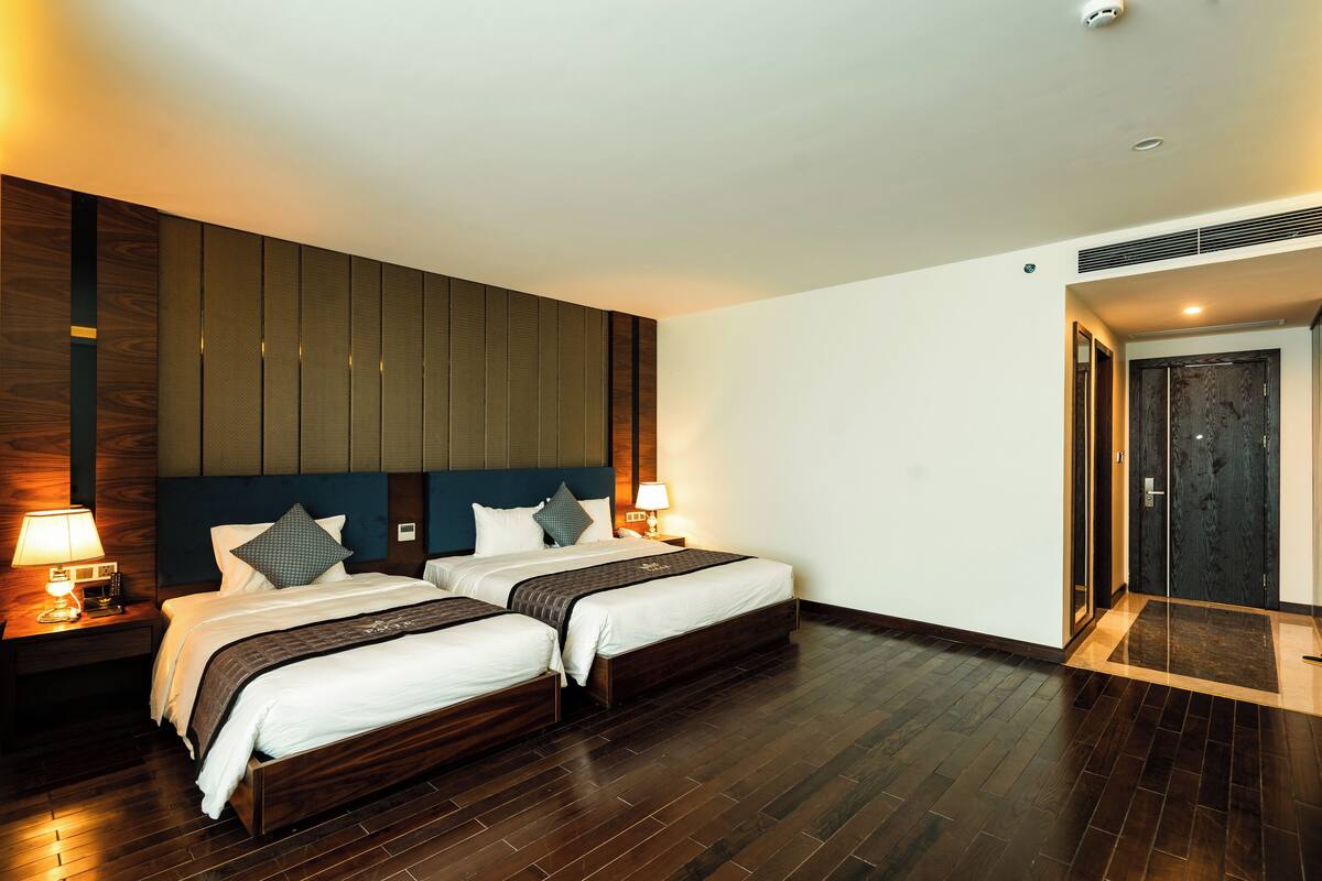 A hotel room with two beds, wooden headboard, dark flooring, and a door with a dark panel.