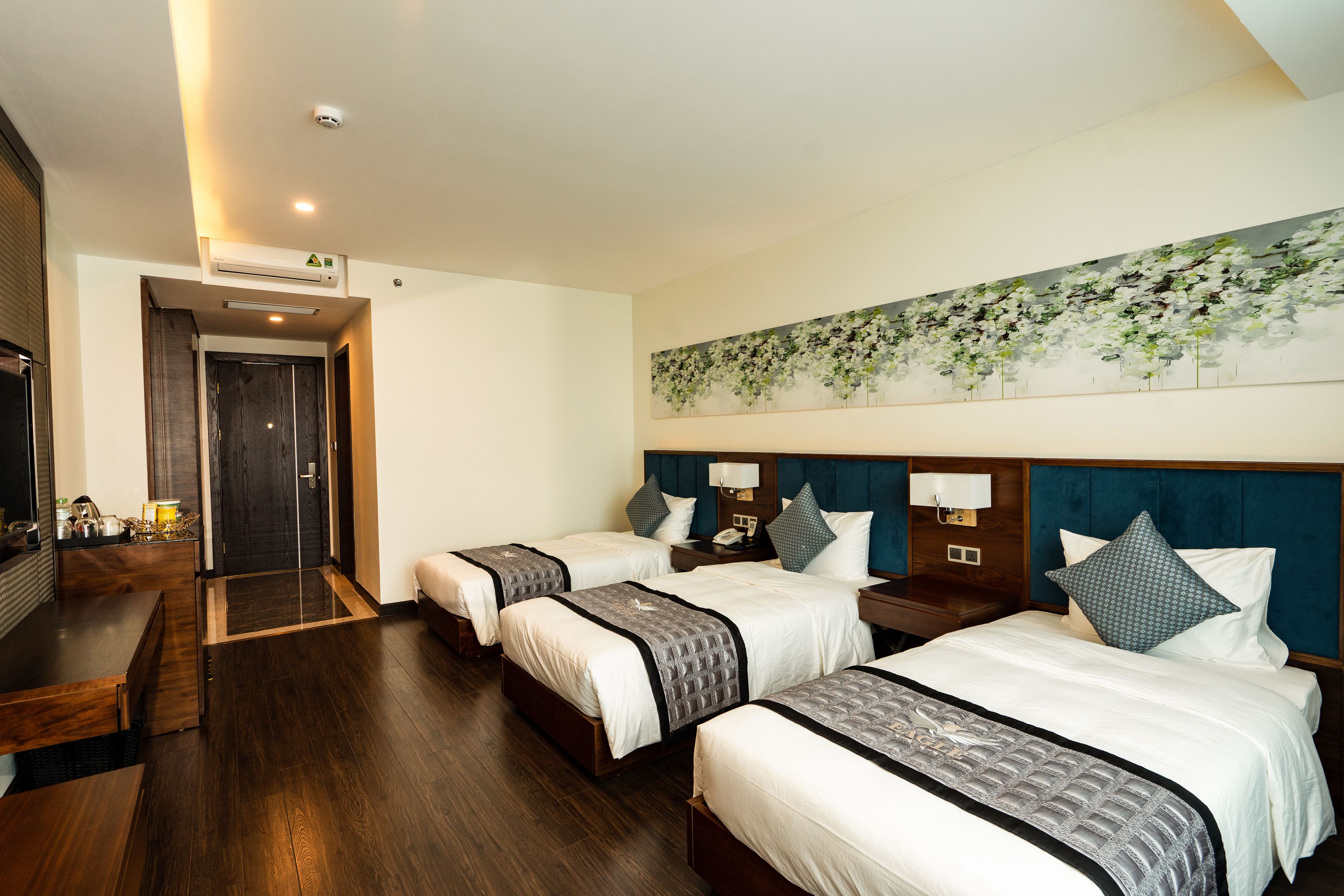 Una habitación de hotel con tres camas, un cuadro de flores grande en la pared, un cabecero de madera y un escritorio pequeño con una lámpara.