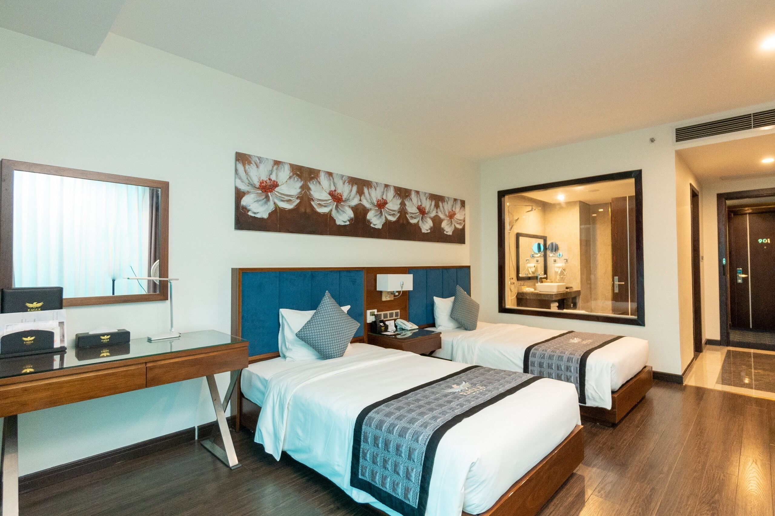 A hotel room with two beds, a desk, a large mirror, and a floral artwork on the wall.