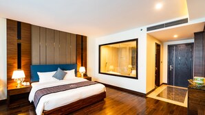 Quarto de hotel moderno com uma cama grande, piso de madeira, cabeceira azul e uma janela ampla.