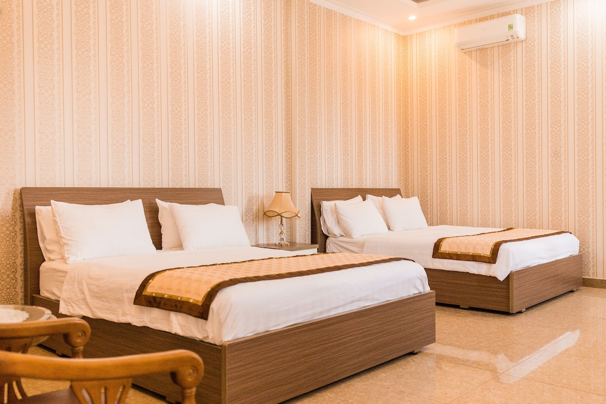 Habitación de hotel con dos camas de madera, ropa de cama blanca, una silla de madera y aire acondicionado montado en la pared.