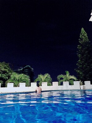 Una piscina nocturna con una persona nadando, rodeada de palmeras y una cerca blanca.
