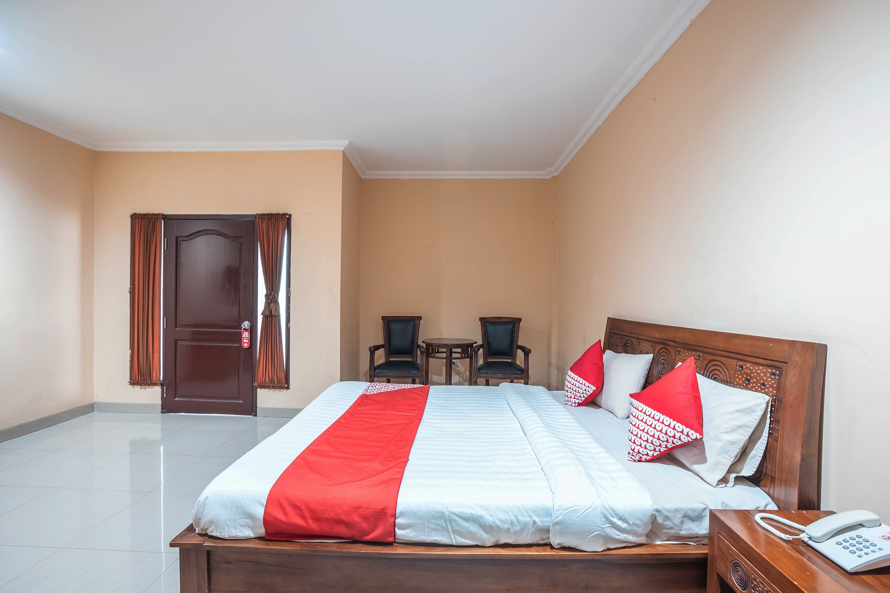 A hotel room with a bed, a bedside table with a phone, a small table with chairs, and a door with curtains.