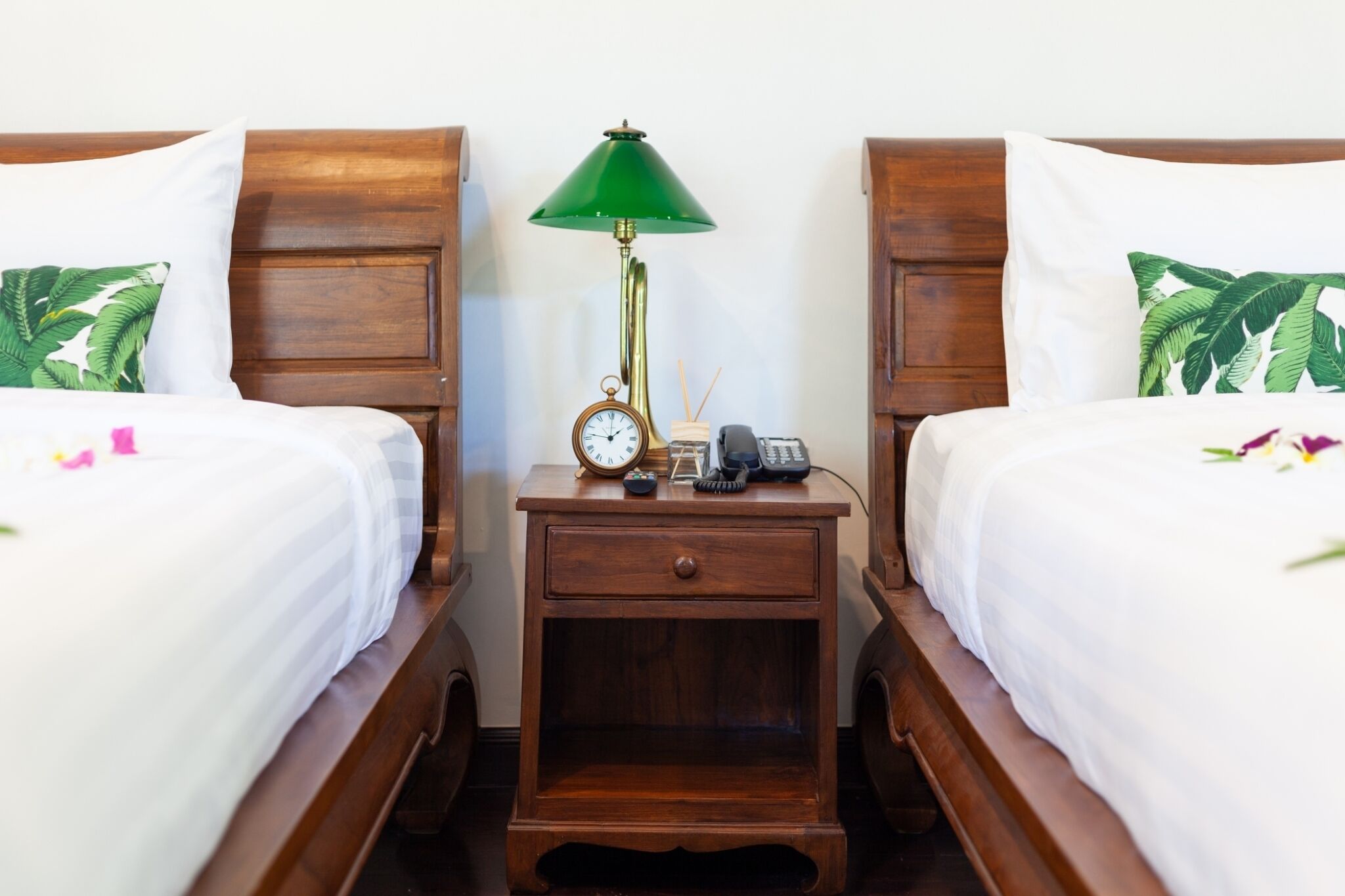 A hotel room with two wooden beds, each with a white bedspread and a pillow with a tropical leaf pattern. There is a wooden nightstand between the beds with a green lamp, a clock, a telephone, and a small decorative item.