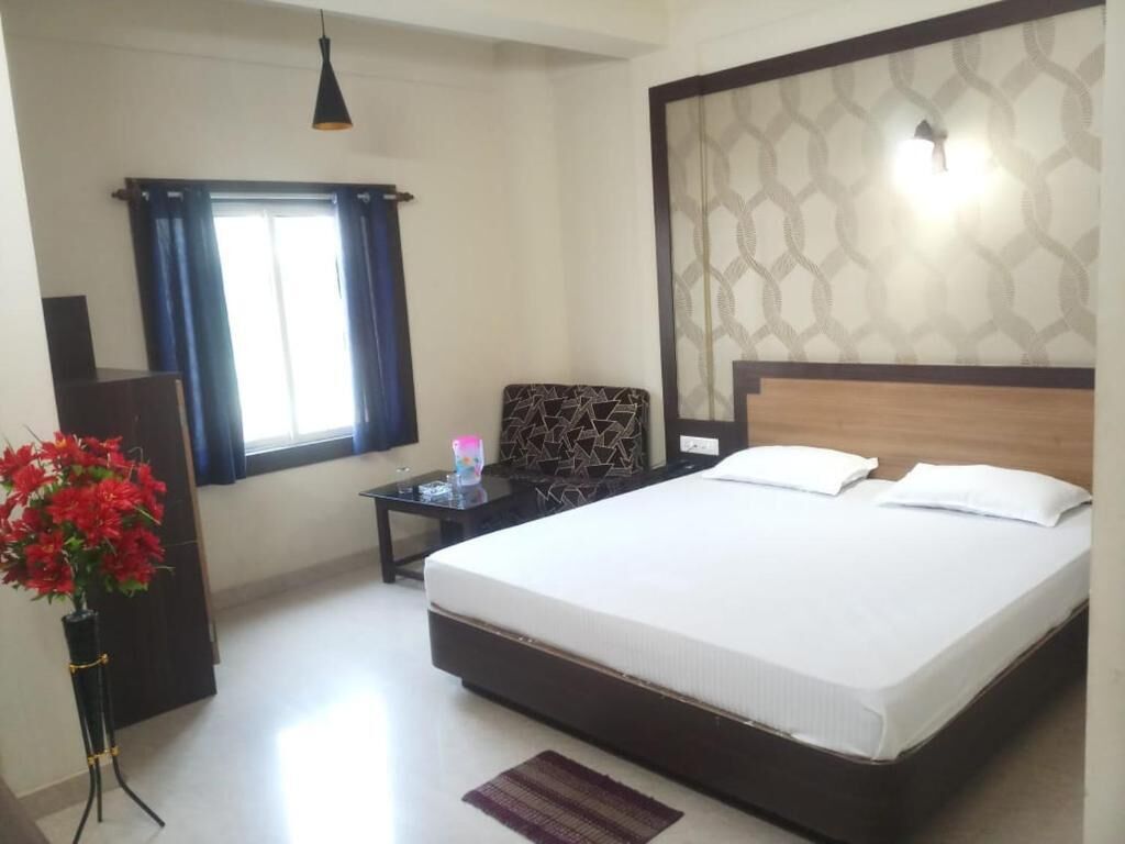 A bedroom with a large bed, a bedside table, a lamp, a window with curtains, and a vase with flowers.