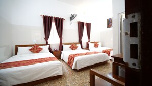 A hotel room with three beds, each with a wooden headboard, white bedding, and red decorative pillows. There is a small bedside table with a lamp, a wall-mounted fan, and a painting on the wall.